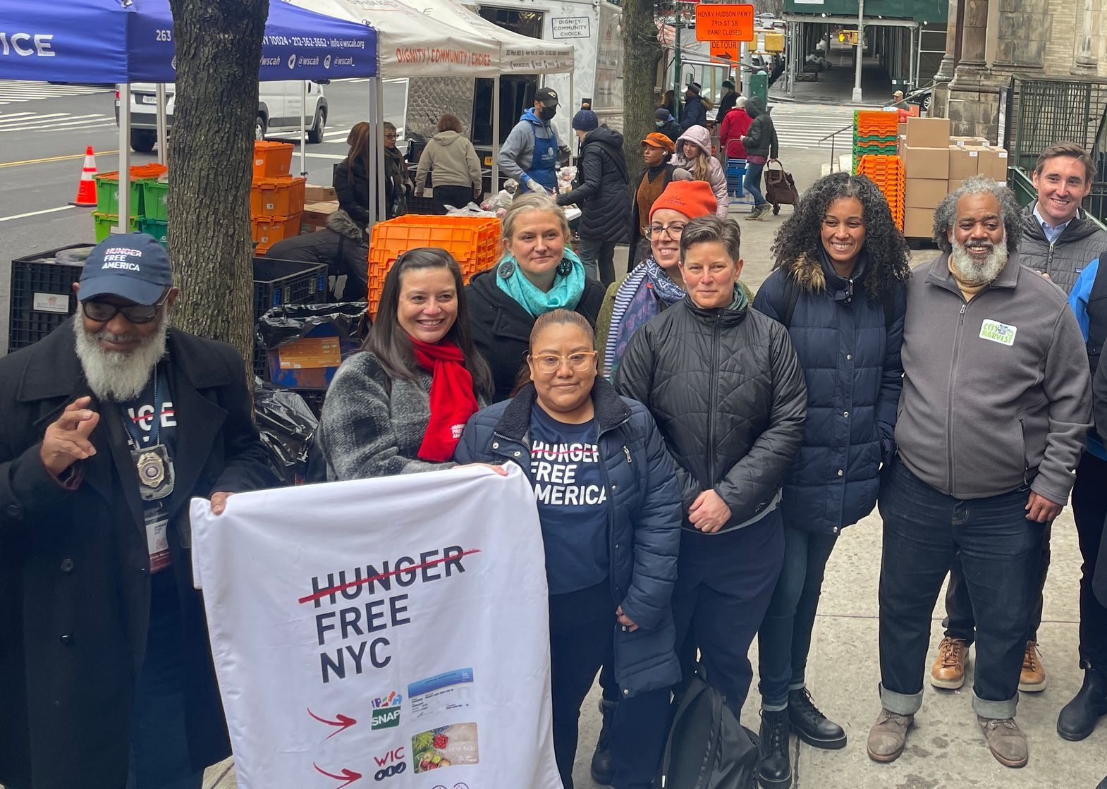 A group of nine advocates stand in front of a WSCAH food distribution site, holding a sign that says "Hunger Free NYC"