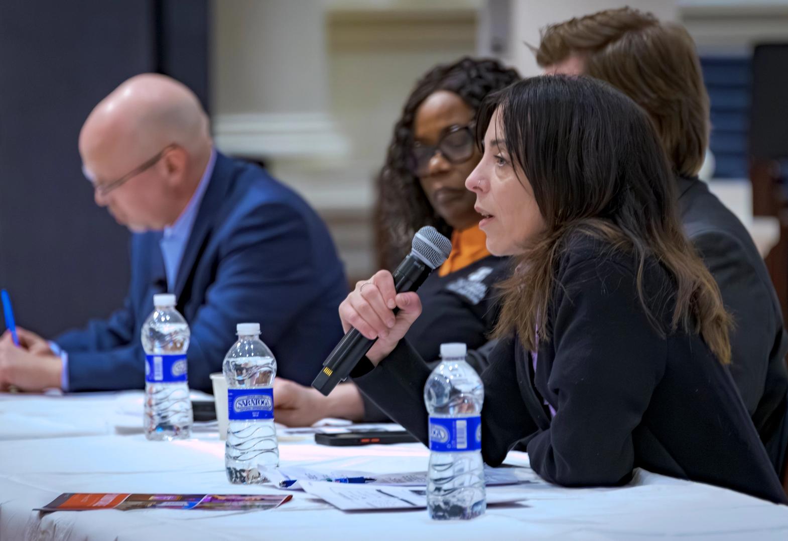 A young woman holding a microphone sits close to the camera, seated at a table with three other people. She is giving testimony at a City Council meeting.