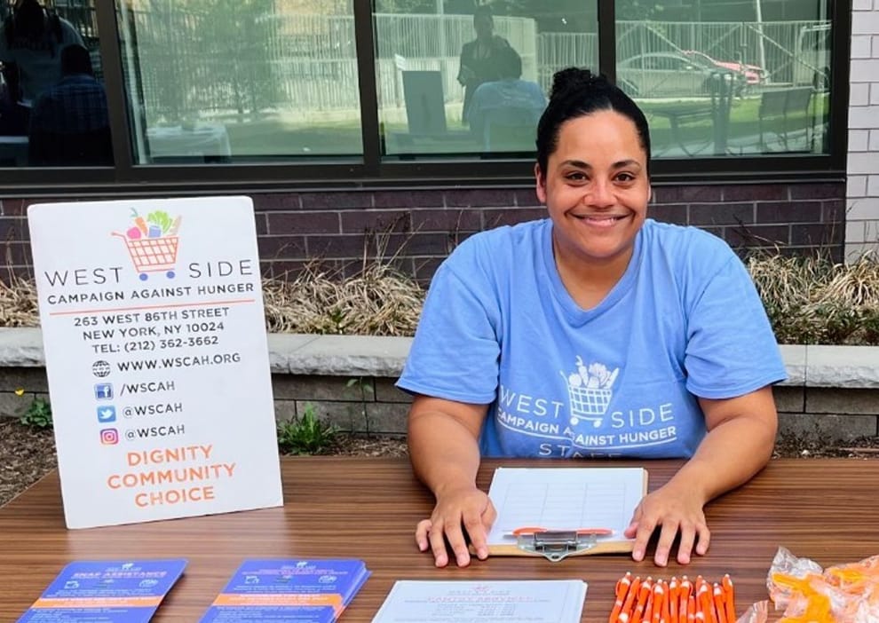 A WSCAH employee sits outside at a table. On the table are handouts, a clipboard where the employee is doing intake, and a placard displaying information about WSCAH.