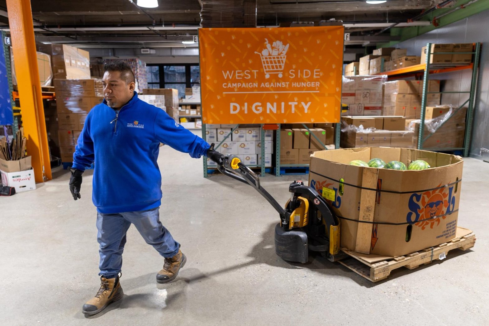 In WSCAH's warehouse, a volunteer pulls a dolly a that carries a large box of watermelons.