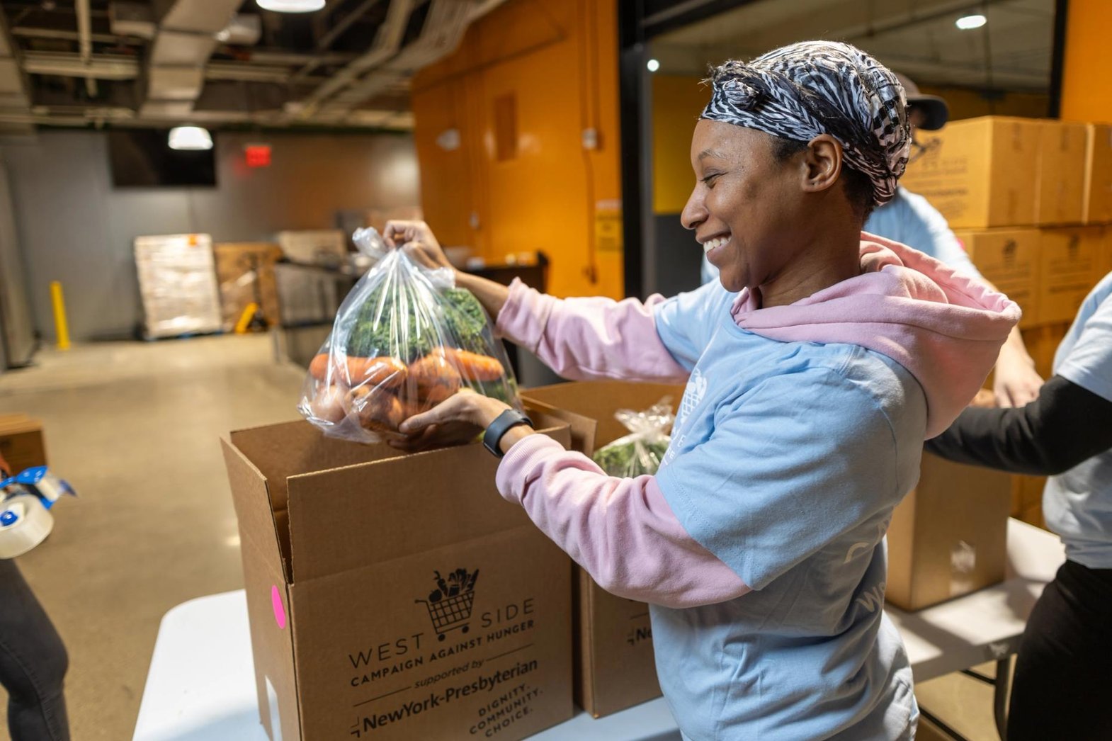 A smiling WSCAH volunteer wearing a bandana places a bag full of fresh produce into a box for distribution.