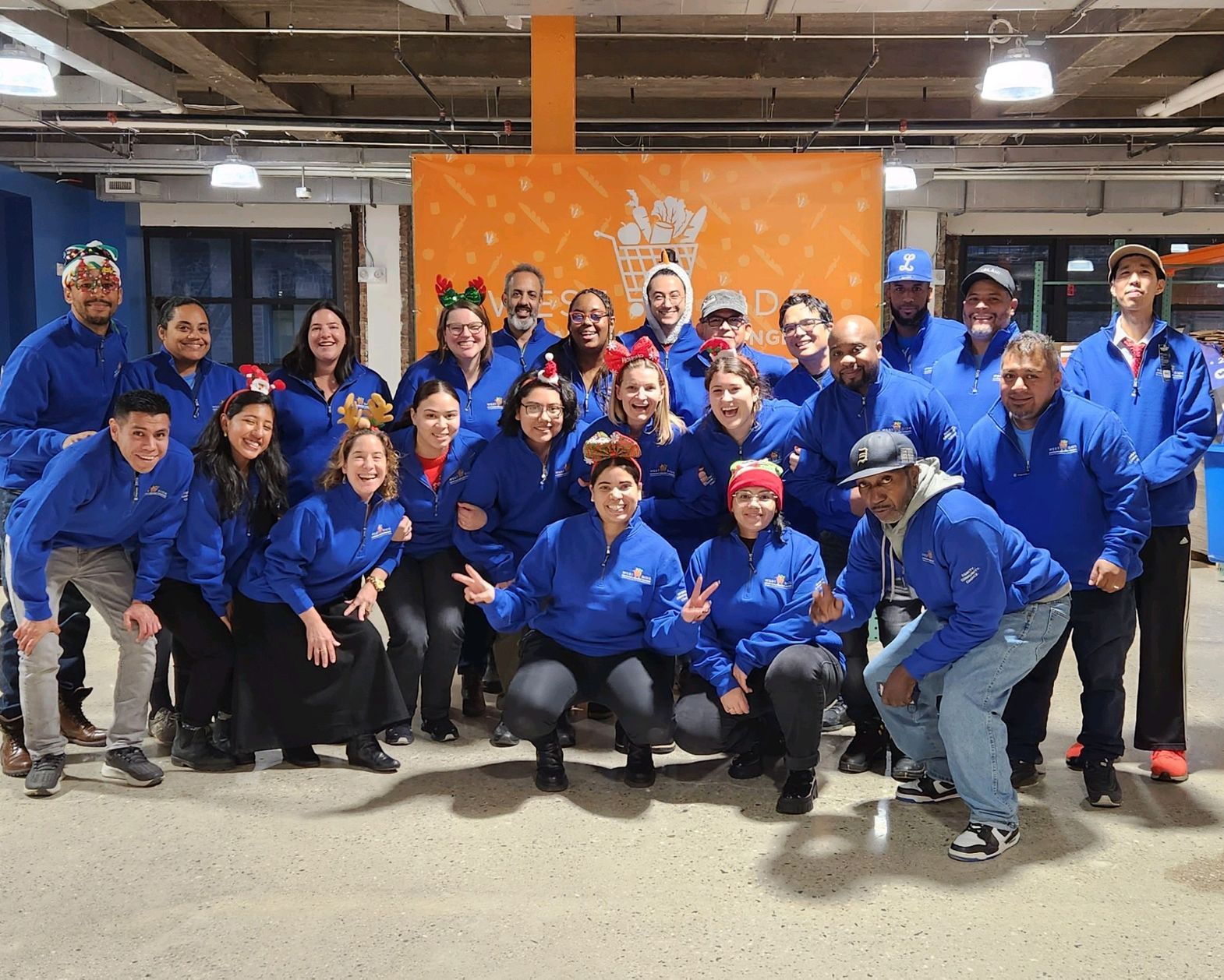 A large group of WSCAH employees stand in our warehouse, smiling, wearing WSCAH-branded jackets and festive holiday adornments such as santa hats.