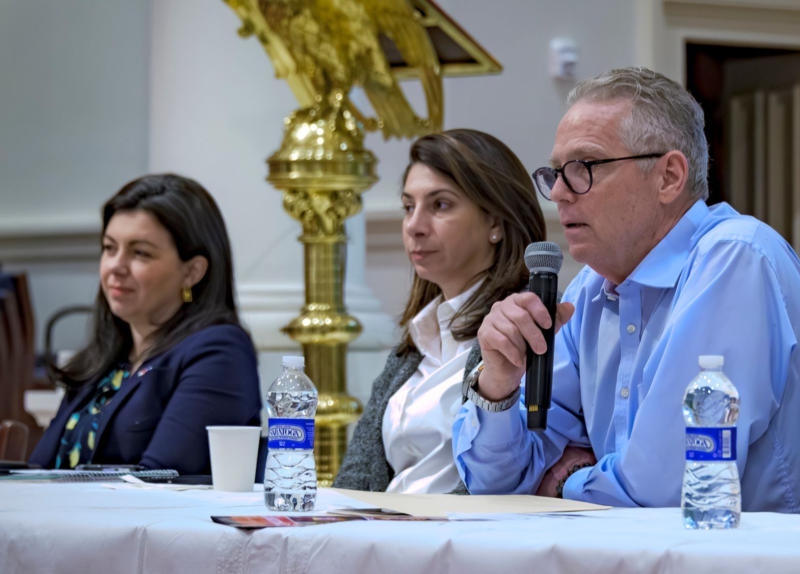 Three of WSCAH's advocates sit at a table in a line, the person closest to the camera is speaking into a microphone. The advocates are at city hall, and behind them is a large, golden statue of an eagle on a column.. 