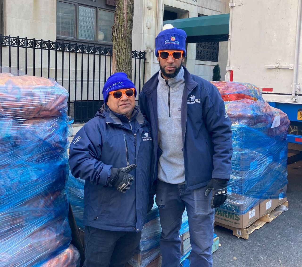 Two members of WSCAH stand in front of palettes stacked with fresh produce, wearing WSCAH-branded hats, sunglasses and jackets.