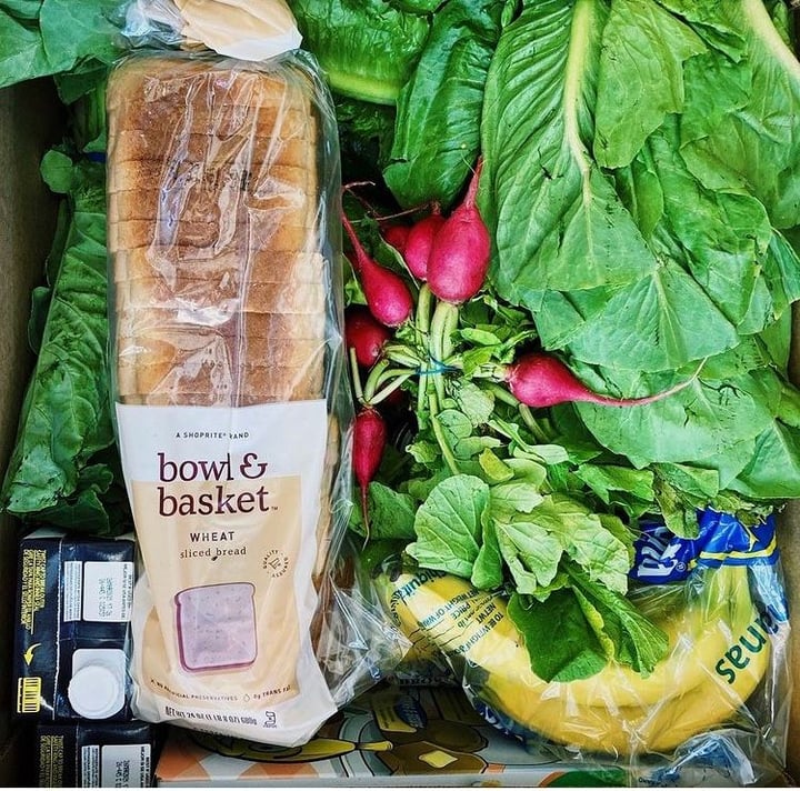 An opened food box. Prominently displayed at the top of the box are a carton of broth, a loaf of sandwich bread, crisp green kale, a bunch of red radishes, and a bag of ripe bananas.