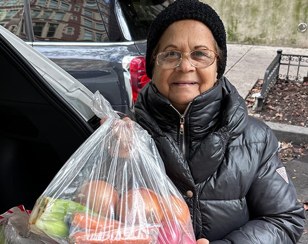 A woman stands in front of her open car trunk, holding a bag of fresh produce.