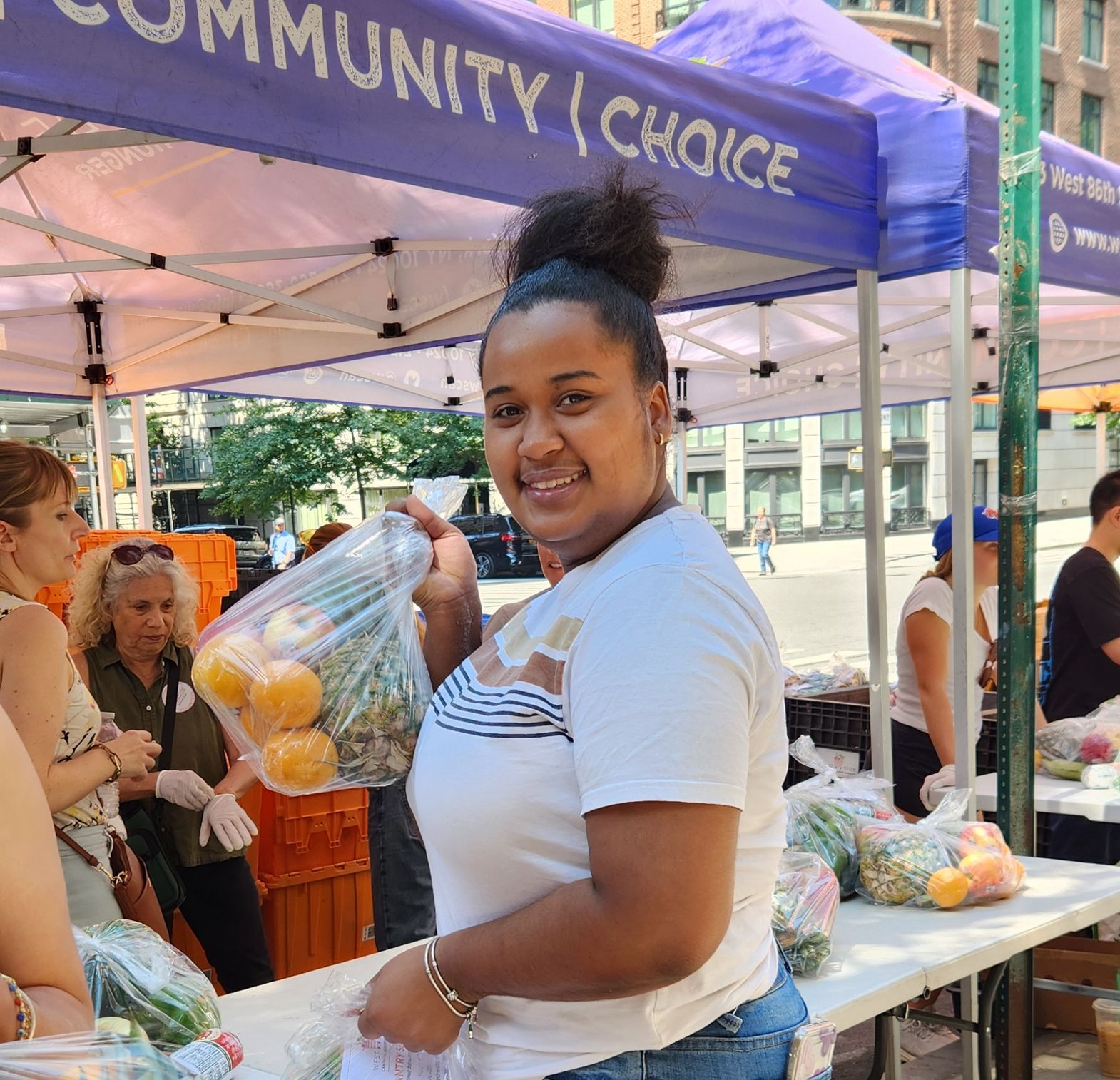A customer smiles over her shoulder at the camera, holding a bag of oranges, apples, and a pineapple. She is stranding in front of a line of WSCAH tents, distributing fruit on a sunny day.