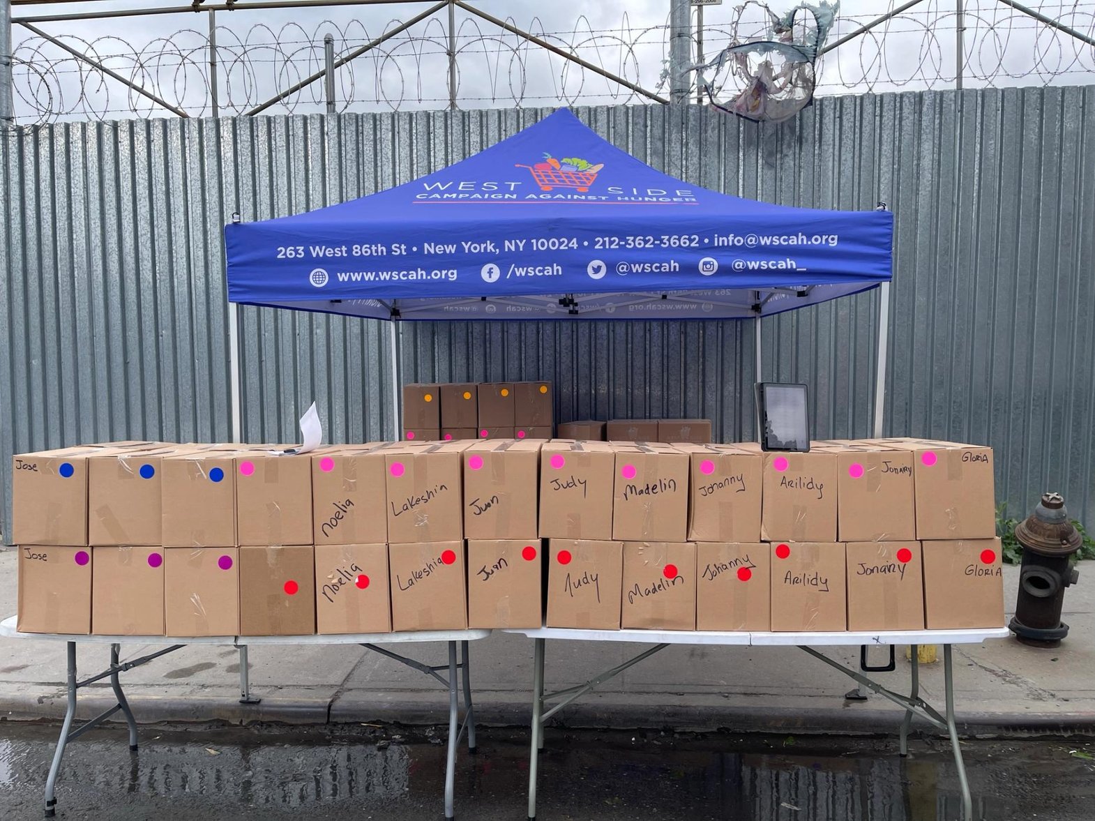 A WSCAH tent is set up on the sidewalk in front of a long metal fence. Closer to the camera, two long plastic tables have a line of food boxes stacked on them, each box labelled with a customer's name.