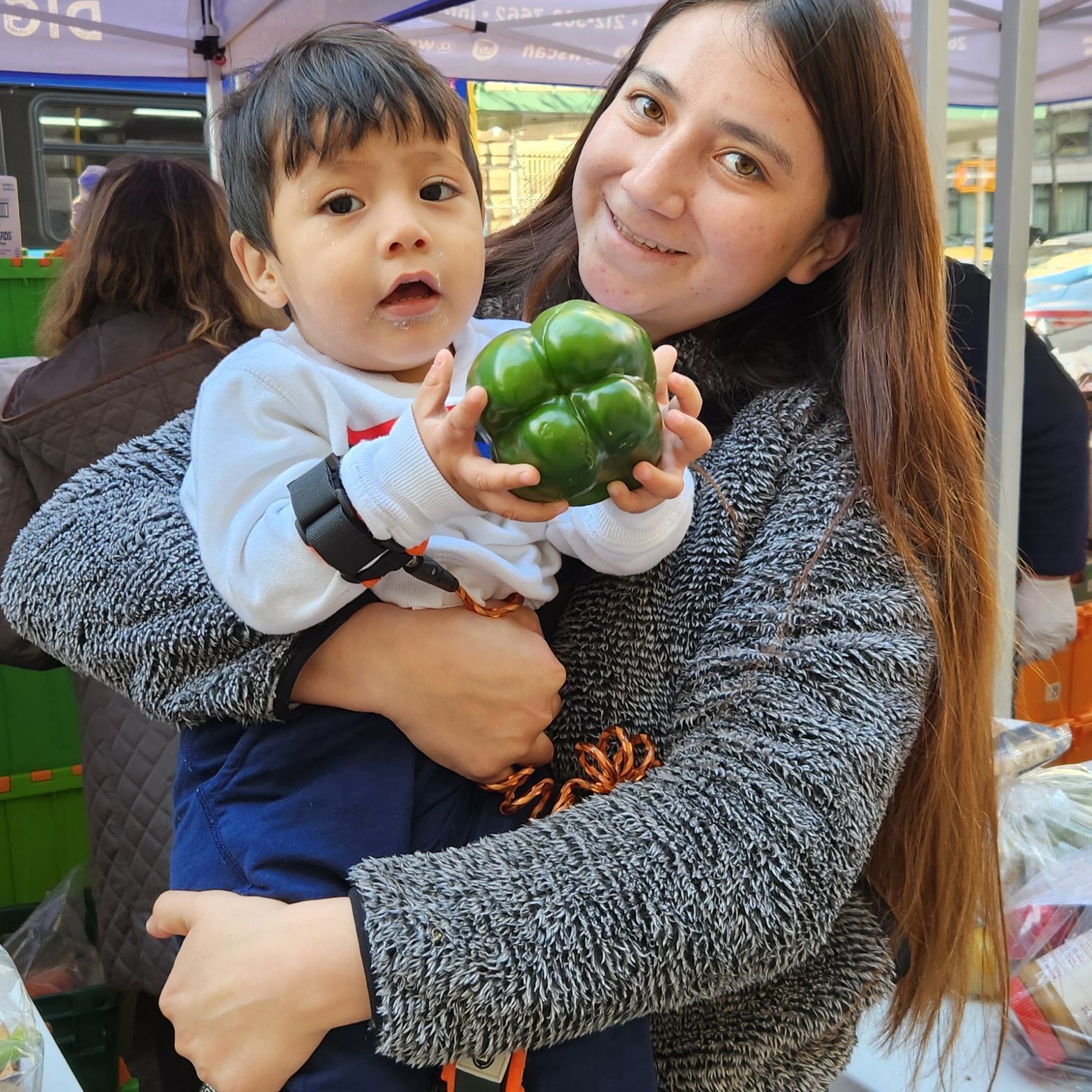 At a WSCAH distribution, a WSCAH customer holds her toddler, who is grasping a green pepper.