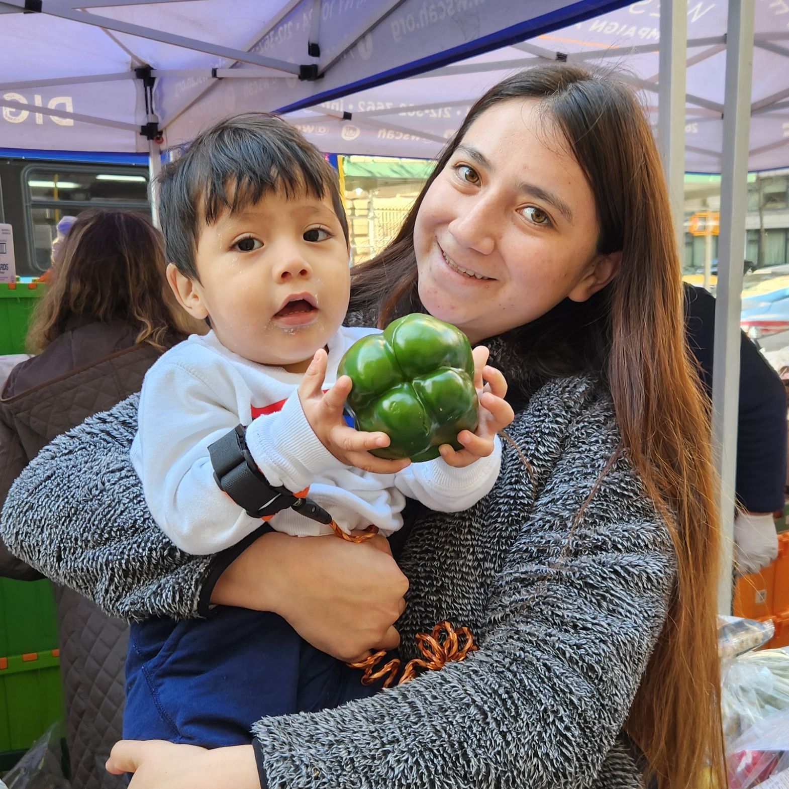 A smiling parent at a WSCAH distribution holds her toddler up to the camera. The child grasps a shiny, green bell pepper.