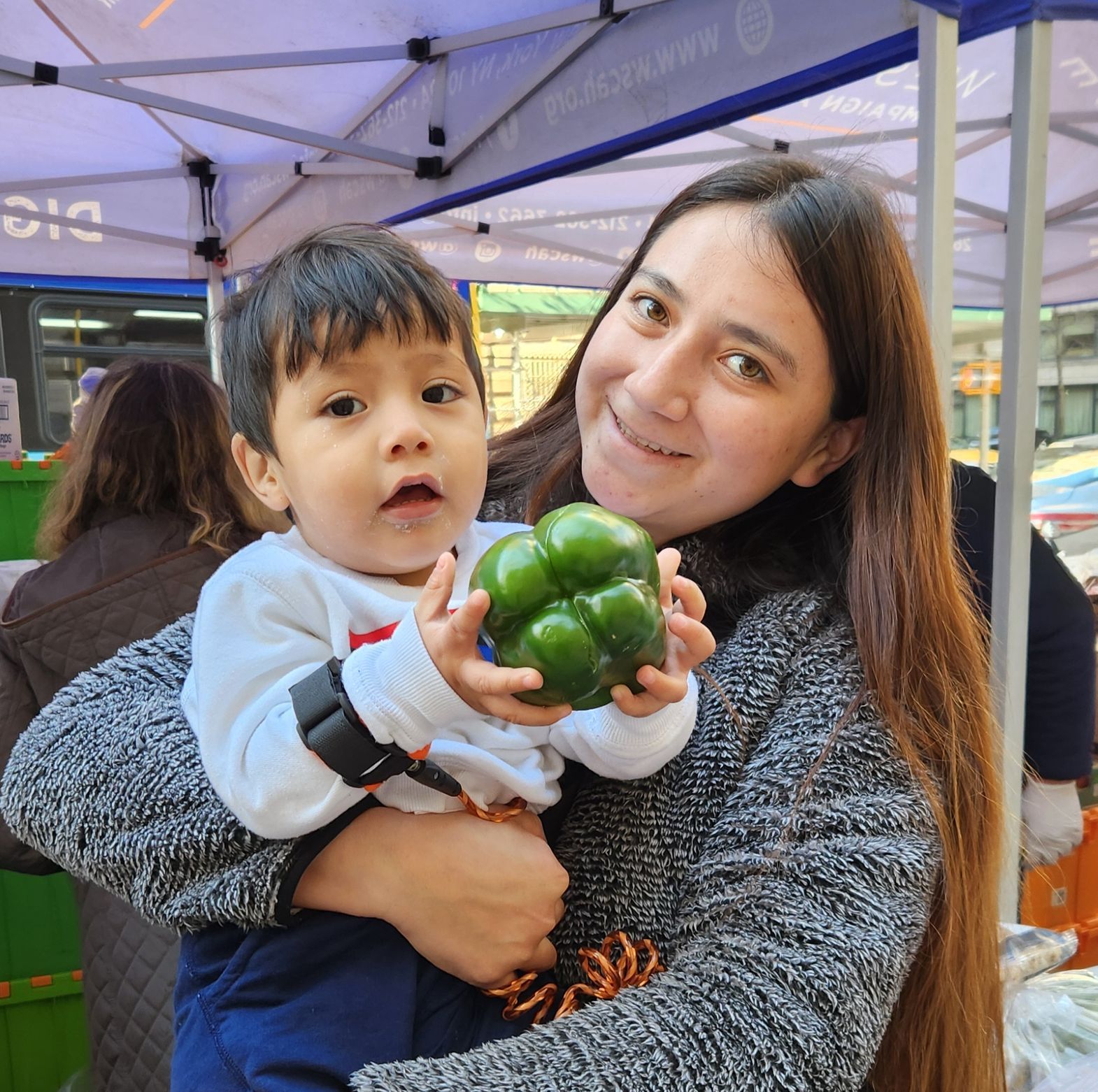 A smiling parent at a WSCAH distribution holds her toddler up to the camera. The child grasps a shiny, green bell pepper.