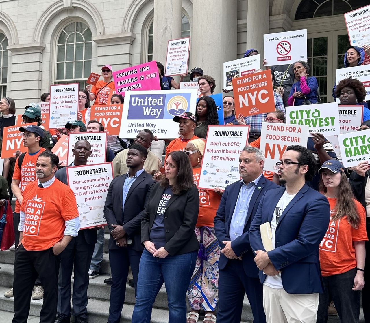 A large group of advocates with The Roundtable, United Way, and City Market stand on the steps of New York City Hall. They hold signs and wear shirts that display slogans like "STAND FOR A FOOD SECURE NYC," "FEEDING NYC FAMILIES IS NOT OPTIONAL," and "Our community is hungry; Cutting CFC will STARVE our pantries."