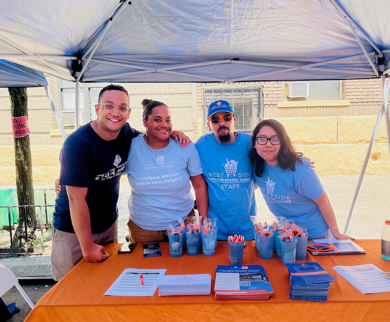 Under a distribution tent, the WSCAH benefits team stands in front of a table displaying pens, papers, flyers, and leaflets for an outreach effort. The ebenfits team is comprised of four smiling people wearing WSCAH-branded shirts, they have their arms around each other's shoulders.