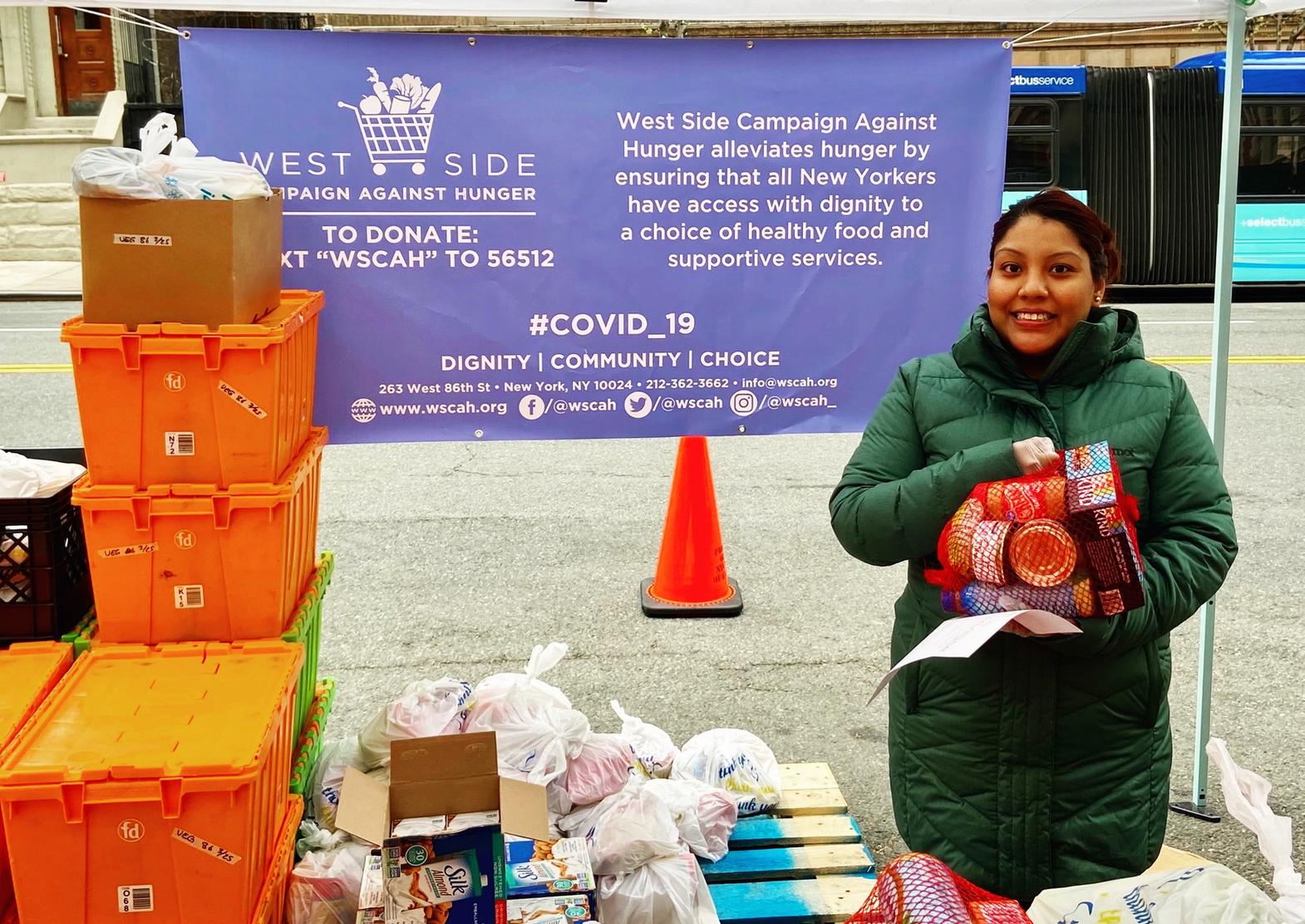 A WSCAH customer wearing a large, green coat holds a stack of packaged food. She is standing inside a WSCAH distribution tent and surrounded by boxes of food.