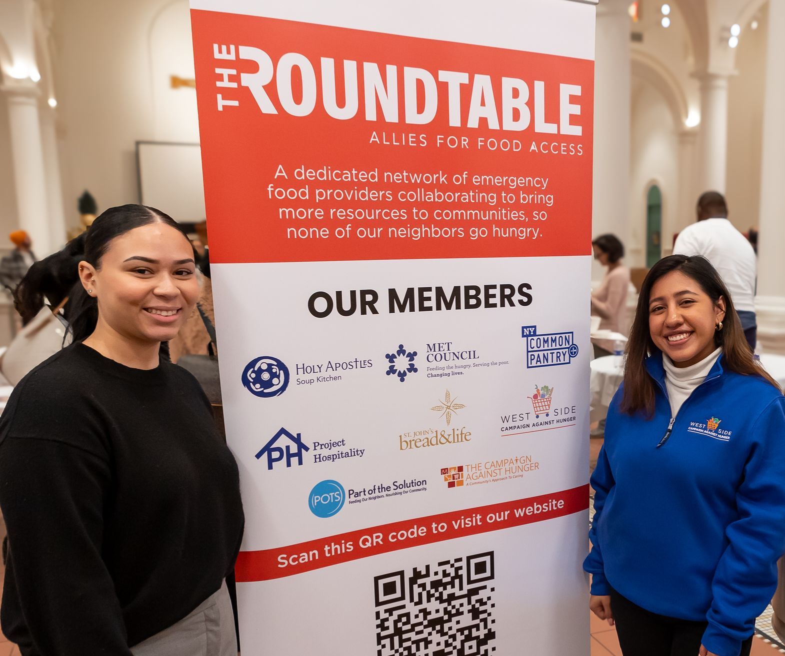 Two members of WSCAH's team stand proudly in front of a sign for "The RoundTable" which lists organizations that are members of the group.