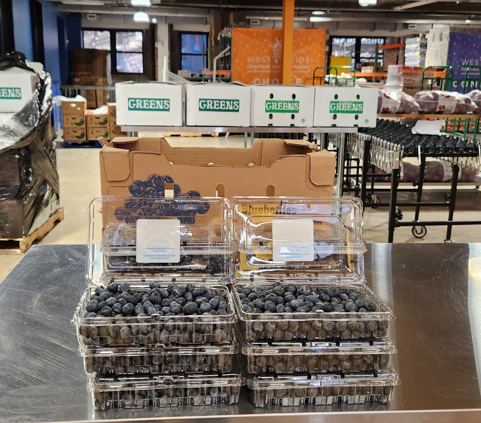 In the WSCAH warehouse, boxes of produce sit stacked on palettes and on steel tables. Six plastic punnets of plump, juicy blueberries sit prominently on a table in front of the camera.